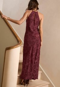 Burgundy maxiklänning med halterneck, texturerat blommönster och nyckelhål bakdetalj, kombinerad med mörka klackskor.