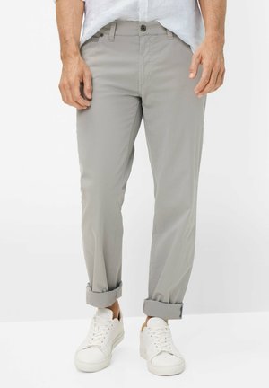 Mann trägt hellgraue hochgekrempelte Chinos und weiße Sneaker, steht vor einem schlichten weißen Hintergrund.