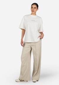 Camiseta blanca de corte holgado con mangas cortas y texto impreso, combinada con pantalones beige de pierna ancha con costuras laterales y un logo.
