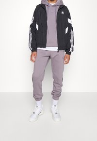 Svart vindjacka med vita ränder, grå hoodie och matchande joggers. Har logotyp, elastiska ärmmuddar och dragsko i botten. Vita sneakers.