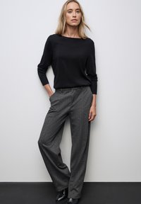 Top negro de punto de manga larga combinado con pantalones de pierna ancha grises que presentan un sutil patrón de espiga. La modelo está de pie sobre una superficie oscura.