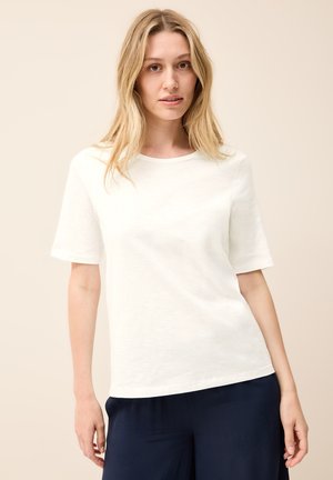 Witte katoenen t-shirt met korte mouwen, ronde hals en een gladde textuur, gecombineerd met donkerblauwe broek, tegen een neutrale achtergrond.