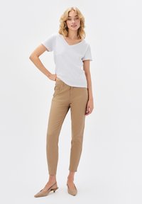 Biała, krótka koszulka typu V z rękawem, zestawiona z jasnobrązowymi dopasowanymi spodniami i beżowymi butami na obcasie, wyeksponowana na jednolitym tle.