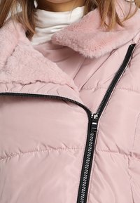 Veste rose matelassée avec col en fausse fourrure, fermeture éclair diagonale et motif de coutures verticales le long du corps, offrant une texture douce.