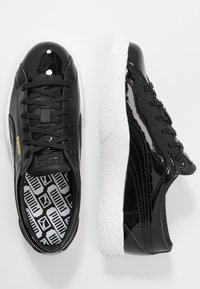 Svarta patenterade lädersneakers med texturerade vita gummisulor, som har svarta snören och ett mönstrat innertyg. Guldaccent med logotyp på sidan.