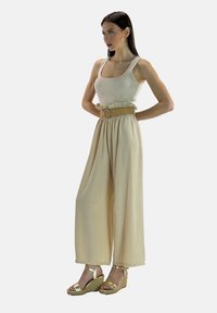 Beige broek met wijde pijpen en geplooide tailleband, gecombineerd met een nauwsluitend licht tanktop. Gouden sleehakken maken de outfit compleet.