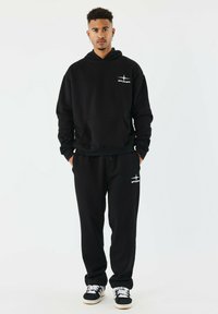 Schwarzer Hoodie mit Fronttasche und Logo, kombiniert mit passenden schwarzen Jogginghosen. Das Outfit bietet eine lockere Passform und weichen Stoff.