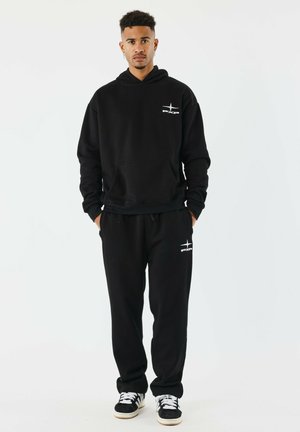 Sudadera negra con bolsillo frontal y logo, combinada con pantalones de chándal negros a juego. El conjunto presenta un corte relajado y una tela suave.