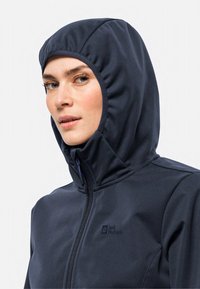 Veste softshell bleu marine avec une capuche ajustée, fermeture éclair à l'avant et logo visible sur la poitrine. Le matériau semble lisse avec une finition mate.