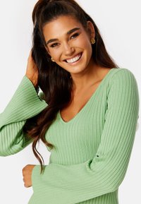 Top verde a costine con scollatura a V, maniche lunghe a campana e silhouette aderente, che mette in mostra una texture morbida e una leggera lucentezza.