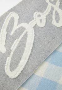 Grå stof med hvid broderet tekst overlapping et lyseblåt og hvidt ternet tekstil. Teksten er iøjnefaldende og tekstureret.