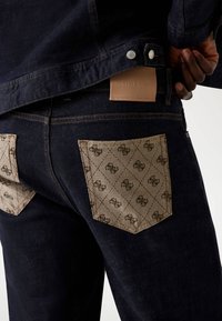 Denim jeans in dark wash with light beige fabric rear pockets featuring a repeating pattern, branded leather tag at the waistband.