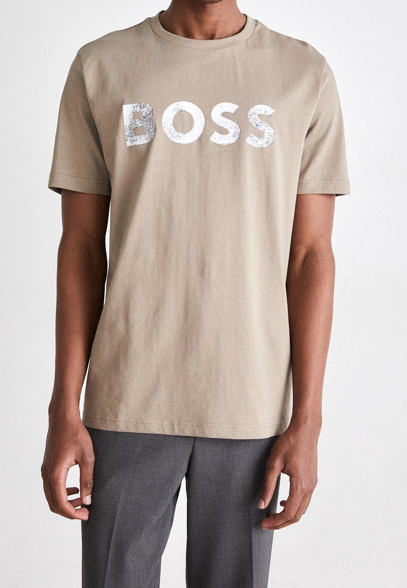 Osoba oblečená v béžovém tričku s bílým třpytivým textem "BOSS" a šedých kalhotách, zobrazená od ramen k polovině stehen na jednolitém pozadí.