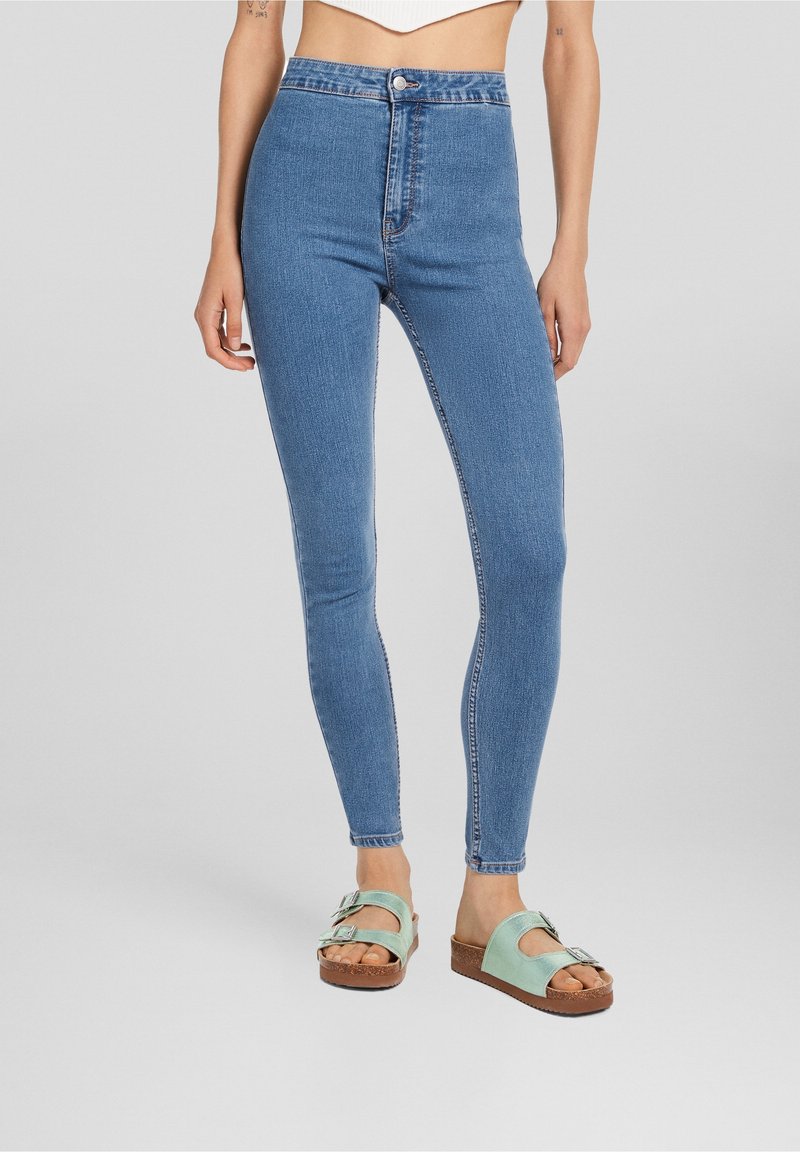 Jean bleu taille haute avec une texture en denim lisse, présentant une fermeture à bouton et des coutures contrastées, associés à des sandales vert menthe.