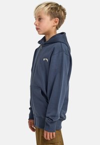 Niebieska bluza z kapturem zapinana na zamek, z regulowanym kapturem, kieszeniami typu kangurka, ściągaczami na rękawach oraz haftowanym logo na piersi. Miękka struktura materiału.