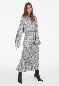 Langes Kleid mit schwarzem und weißem Schlangenprint, einem kragenförmigen Ausschnitt und weiten Ärmeln. Getragen mit schwarzen Knöchelstiefeln. Glatte Stofftextur.
