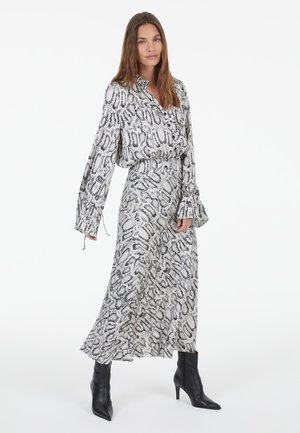 Langes Kleid mit schwarzem und weißem Schlangenprint, einem kragenförmigen Ausschnitt und weiten Ärmeln. Getragen mit schwarzen Knöchelstiefeln. Glatte Stofftextur.