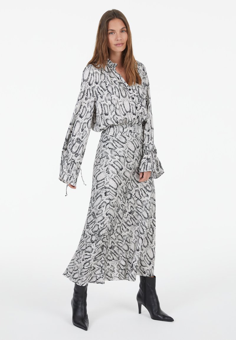 Langes Kleid mit schwarzem und weißem Schlangenprint, einem kragenförmigen Ausschnitt und weiten Ärmeln. Getragen mit schwarzen Knöchelstiefeln. Glatte Stofftextur.