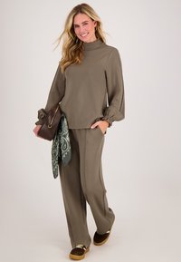 Vrouw met lang blond haar, gekleed in een olijfgroen, losvallend top en broek, met een bruine handtas met een groen patroon sjaal, lachend.