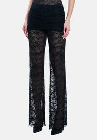 Modella che indossa pantaloni trasparenti in pizzo floreale nero con una minigonna aderente sopra e tacchi neri con punta a mandorla su sfondo bianco.