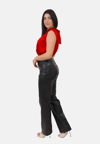 Donna con lunghi capelli scuri che indossa un top senza maniche rosso, pantaloni di pelle nera e sandali con tacco alto bianchi, che posa di lato.