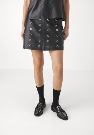 Partie inférieure du corps vêtue d'une jupe en cuir noire et d'un haut avec œillets argentés, chaussettes noires et mocassins noirs avec clous argentés sur fond blanc.