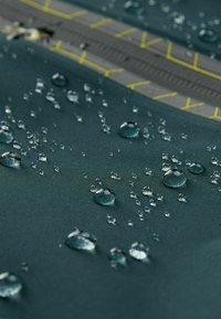 Tissu imperméable de couleur vert foncé avec des gouttes d'eau qui perlaient à la surface. Présente une fermeture à glissière grise avec des accents de surpiqûres jaunes.