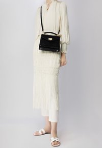 Abito bianco textured con orlo a frange, abbinato a una borsa a tracolla nera con effetto coccodrillo e sandali bianchi flat con design a strap.