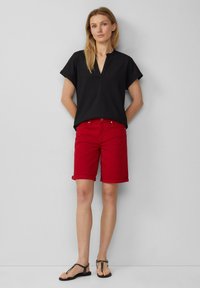 Zwart topje met korte mouwen en een V-hals, gecombineerd met felrode knielange shorts. De shorts hebben een omgeslagen zoom. Zwarte platte sandalen maken de look af.