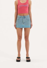 Rosa, gekürztes Tanktop mit gesticktem Text, kombiniert mit einem hellblauen Denim-Minirock mit blauer Schrift auf der Vorderseite. Schwarze Plateausandalen.