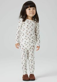 Ensemble de pyjama blanc à manches longues arborant un motif de nœuds marron répété. Matière en coton doux avec un pantalon ajusté. Chaussures à enfiler marron.