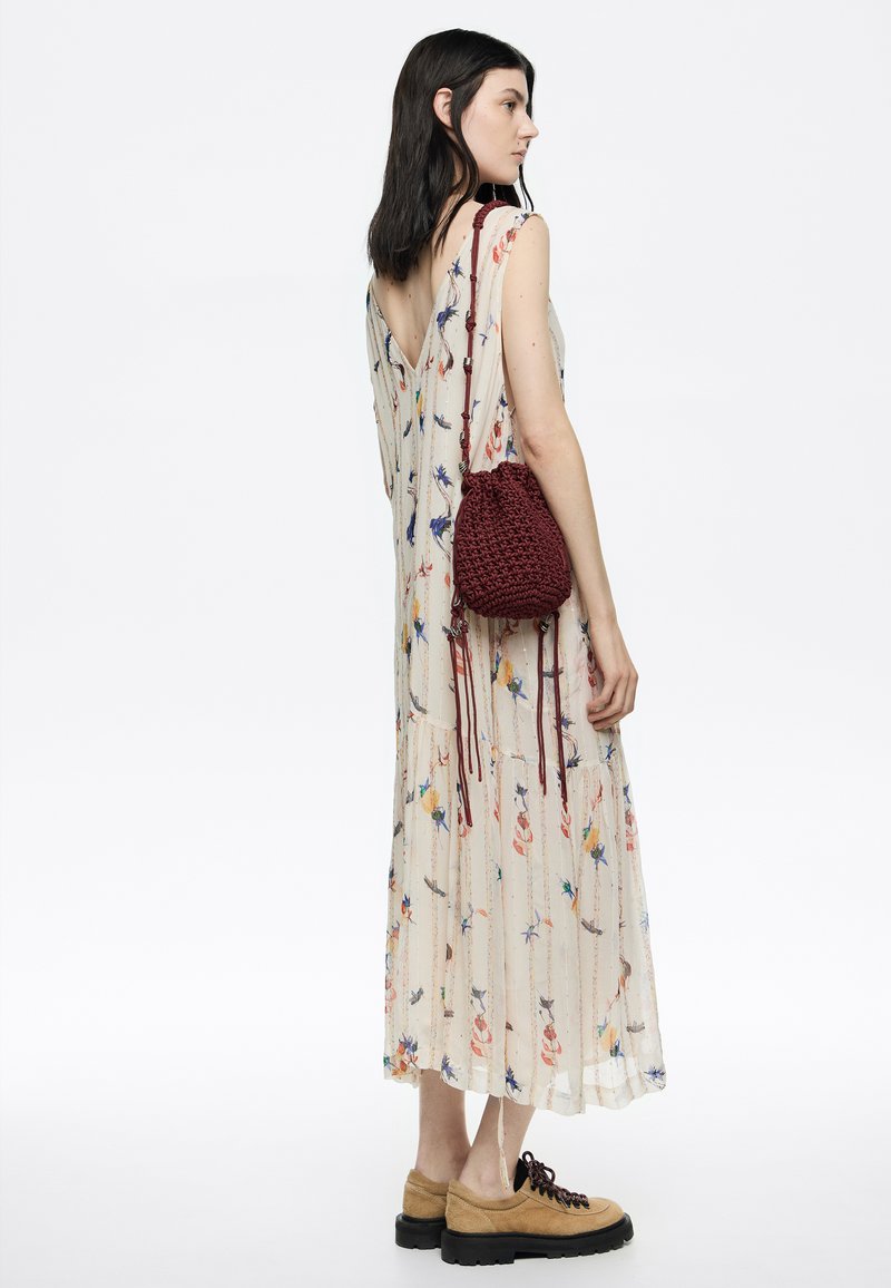 Vestido de crema, transparente y con estampado floral, con espalda en V, combinado con un bolso de ganchillo color marrón y zapatos beige con suelas negras y cordones oscuros.