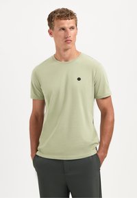 Man met kort, krullend haar, draagt een lichtgroen T-shirt met korte mouwen met een klein zwart hexagon logo, handen in de zakken, kijkt opzij.