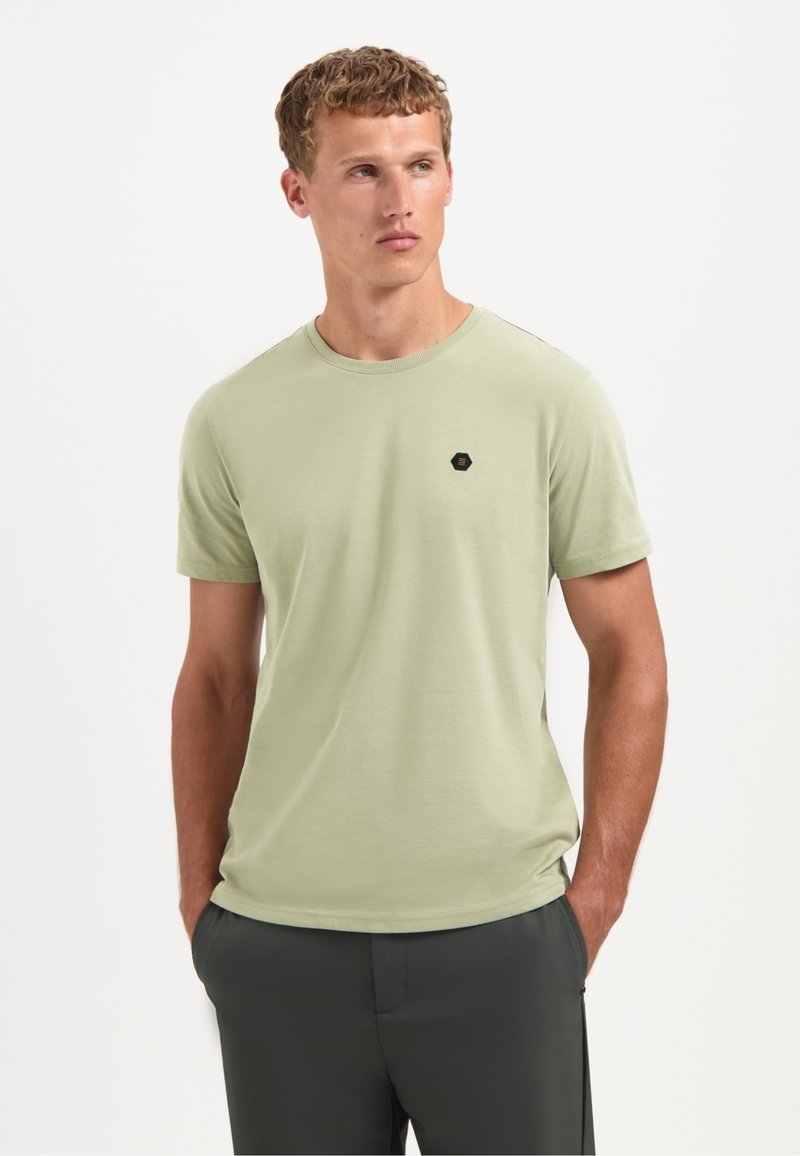 Man met kort, krullend haar, draagt een lichtgroen T-shirt met korte mouwen met een klein zwart hexagon logo, handen in de zakken, kijkt opzij.