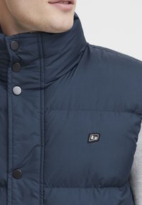 Temno modra napihnjena brezrokavnika z visokim ovratnikom, črnimi pritrdilnimi gumbi in logotipom na prsih. Ima kviltano teksturo in trpežen material.