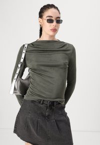 Top lungo a maniche con scollo drappeggiato in verde oliva, design aderente, abbinato a una mini gonna in denim nera e una borsa argentata.
