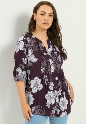 Frau, die eine dunkelviolette Bluse mit großen weißen Blumenmustern und hochgekrempelten Ärmeln trägt, kombiniert mit hellblauen Jeans, steht vor einer weißen Wand.