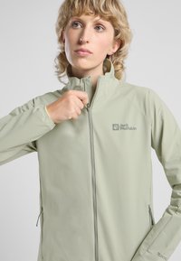 Giacca verde chiaro con zip completa e colletto alto, realizzata in tessuto morbido e traspirante. Presenta tasche laterali e un logo sul lato sinistro del petto.