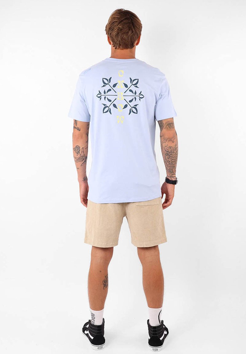 T-shirt bleu clair à manches courtes avec une impression graphique en bleu foncé et jaune au dos, associé à un short beige.