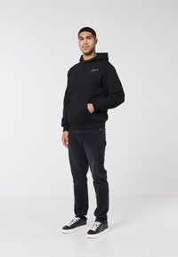 Mann steht mit der Hand in der Tasche, trägt einen schwarzen Hoodie, schwarze Jeans und schwarze Sneakers mit weißen Sohlen und Schnürsenkeln vor einem schlichten weißen Hintergrund.