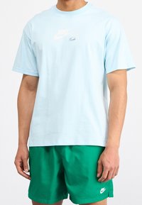 Světle modré bavlněné tričko s kulatým výstřihem, s bílým logem Nike a malým iridescentním detailem na přední straně, nošeno s zelenými šortkami.