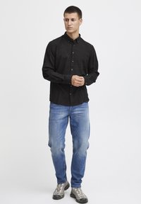 Camisa negra de botones con cuello, tela texturizada, herrajes dorados, combinada con jeans de mezclilla azul claro y zapatillas deportivas grises.
