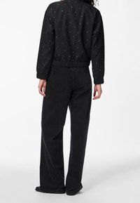 Svart kort jacka med silverfärgade prickar, elastiska ärmslut och midja, tillsammans med svarta vida jeans, visad bakifrån.