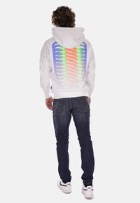 Felpa bianca con un grafico a forma di colonna vertebrale colorato in tonalità arcobaleno sulla schiena, abbinata a jeans blu scuro e sneakers bianche.