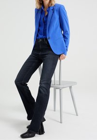 Blå kavaj med hakkragar över en blå skjorta med knappar, kombinerat med mörka denimjeans och svarta stövlar med klack. Enkel, modern design.