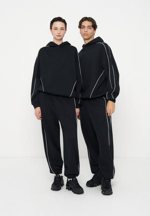 Due persone stanno fianco a fianco indossando felpe con cappuccio nere abbinate e pantaloni con profili bianchi e sneakers nere chunky su uno sfondo semplice.