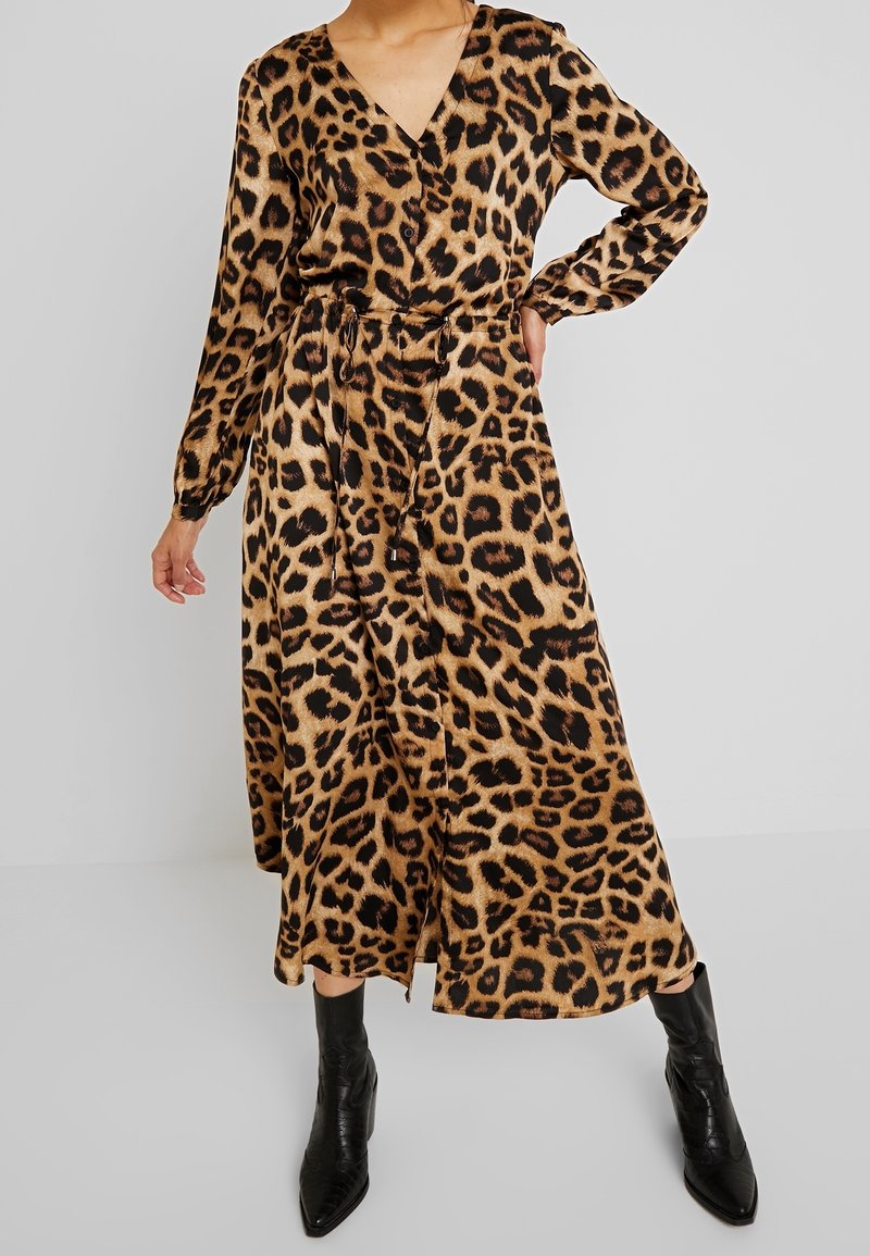 Femme portant une robe midi à manches longues imprimée léopard avec une ceinture nouée à la taille et des bottines noires à bout pointu, sur un fond gris uni.