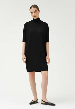 Czarna sukienka z krótkim rękawem i kołnierzem typu mock turtleneck, wykonana z gładkiego materiału. Model nosi czarne buty wsuwane. Prosty, minimalistyczny design.