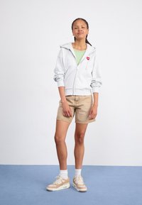 Grauer Zip-Hoodie mit einem roten Herzpatch, darunter ein grünes Oberteil, beige Shorts und beige Sneakers. Vor einem schlichten Hintergrund.