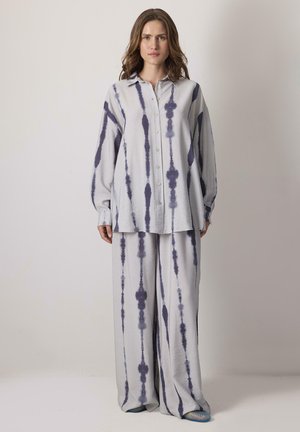 Nő áll, laza gombos ingben és széles szárú nadrágban, amely világos anyagon sötét, függőleges batikcsíkos mintázattal rendelkezik, egyszínű háttér előtt.